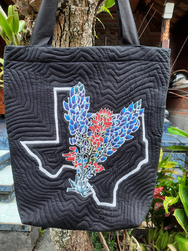 A handpainted batik, lightly quilted shoulder bag with a black background. A beautifully intricate design of Bluebonnets and Indian paintbrush flowers in a bouquet are laid over the Texas boundary.