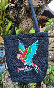 Handpainted batik shoulder bag with a stylized macaw with its wings stretched up, perched on a short branch. Below the bird are the words "parrot addict" also batiked & painted by hand. The background color is black and the bag is lightly quilted to give texture and a bit of a 3D effect.