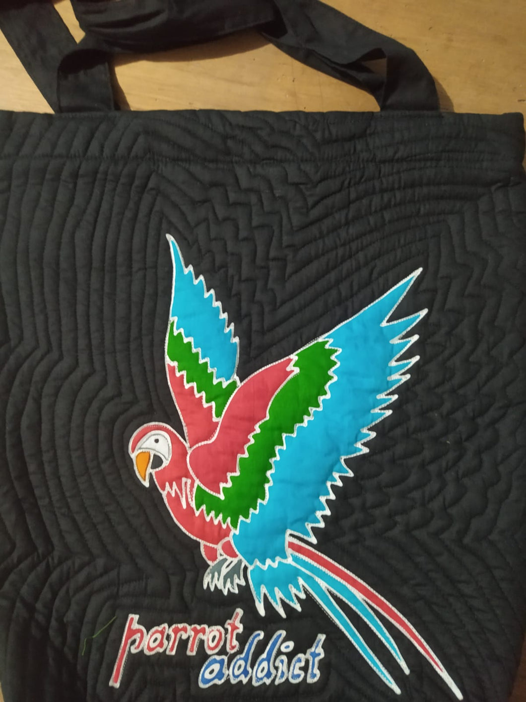 A stylized macaw with outspread wings has been batiked and painted by hand onto this quilted should bag. The words 