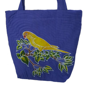 A yellow Indian Ringneck parakeet is perched on lush tropical foliage. Background color is blue/violet color.  The bag's design is batiked and dyed by hand.