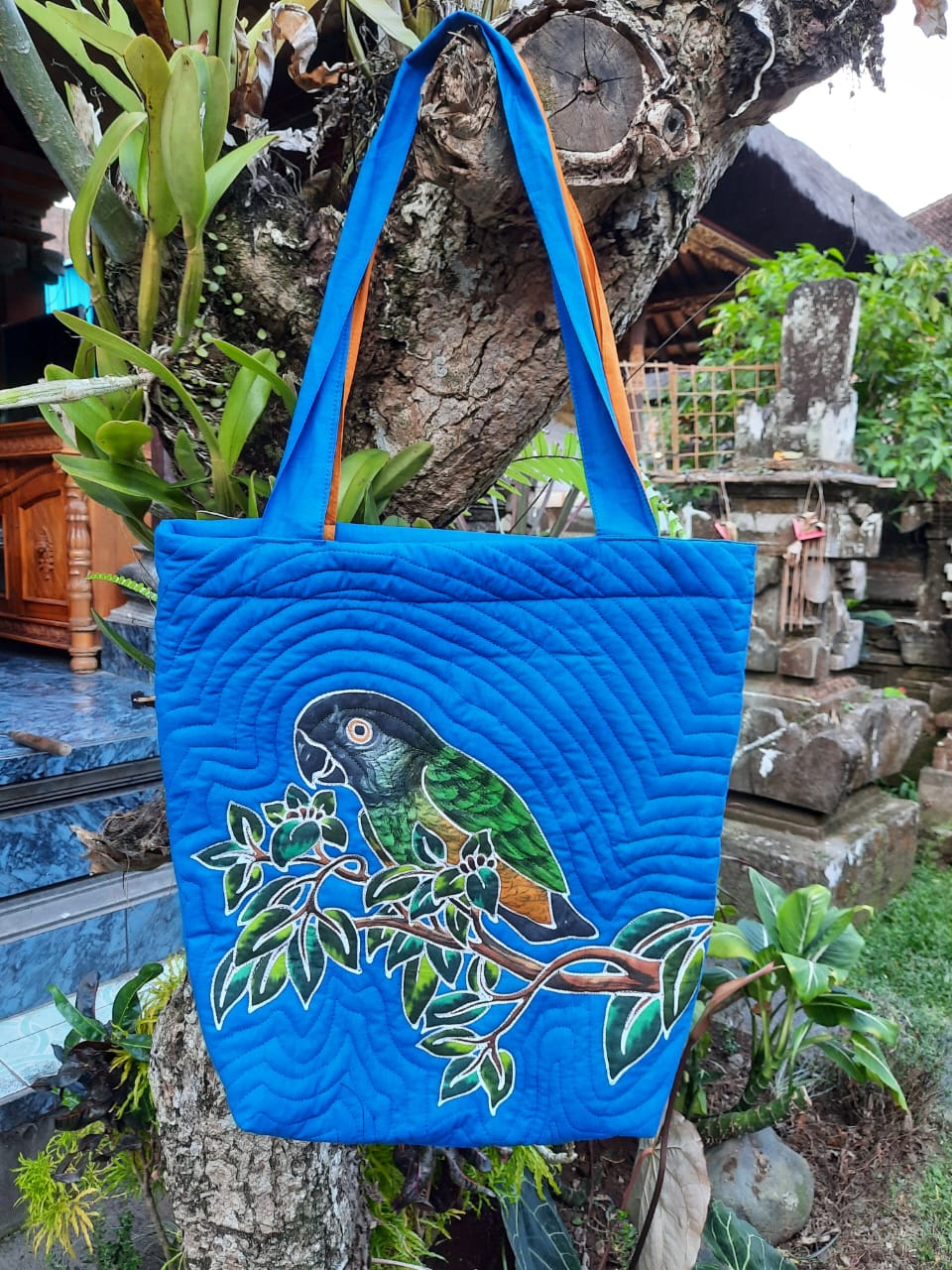 A handpainted batik shoulder bag featuring the popular Senegal Parrot. The bird is perched on a leafy branch. The background color is blue and the quilting adds textural detail to the design.