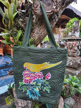 A shoulder bag featuring a handpainted batik yellow Indian Ringneck parakeet set among red & yellow tropical flowers and foliage. The black color is a very dark green.
