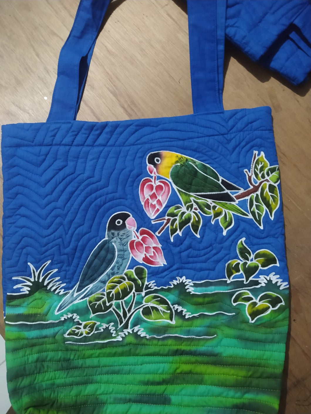 A normal Black-masked lovebird and a blue mutation each hold a heart-shaped red & white flower in their beak. One is perched on a branch, the other standing on varied green groundcover. This handpainted batik bag has blue shoulder straps matching the color of the sky.