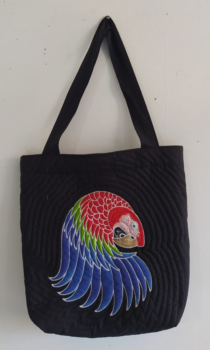 A hand-painted batik tote-style bag featuring a stylized Greenwing macaw against a black background.